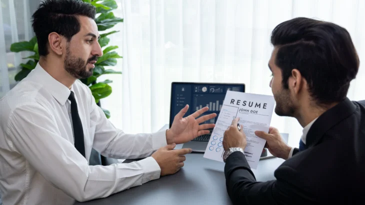 Two men sit at a desk, one holding a resume, likely in a job interview setting